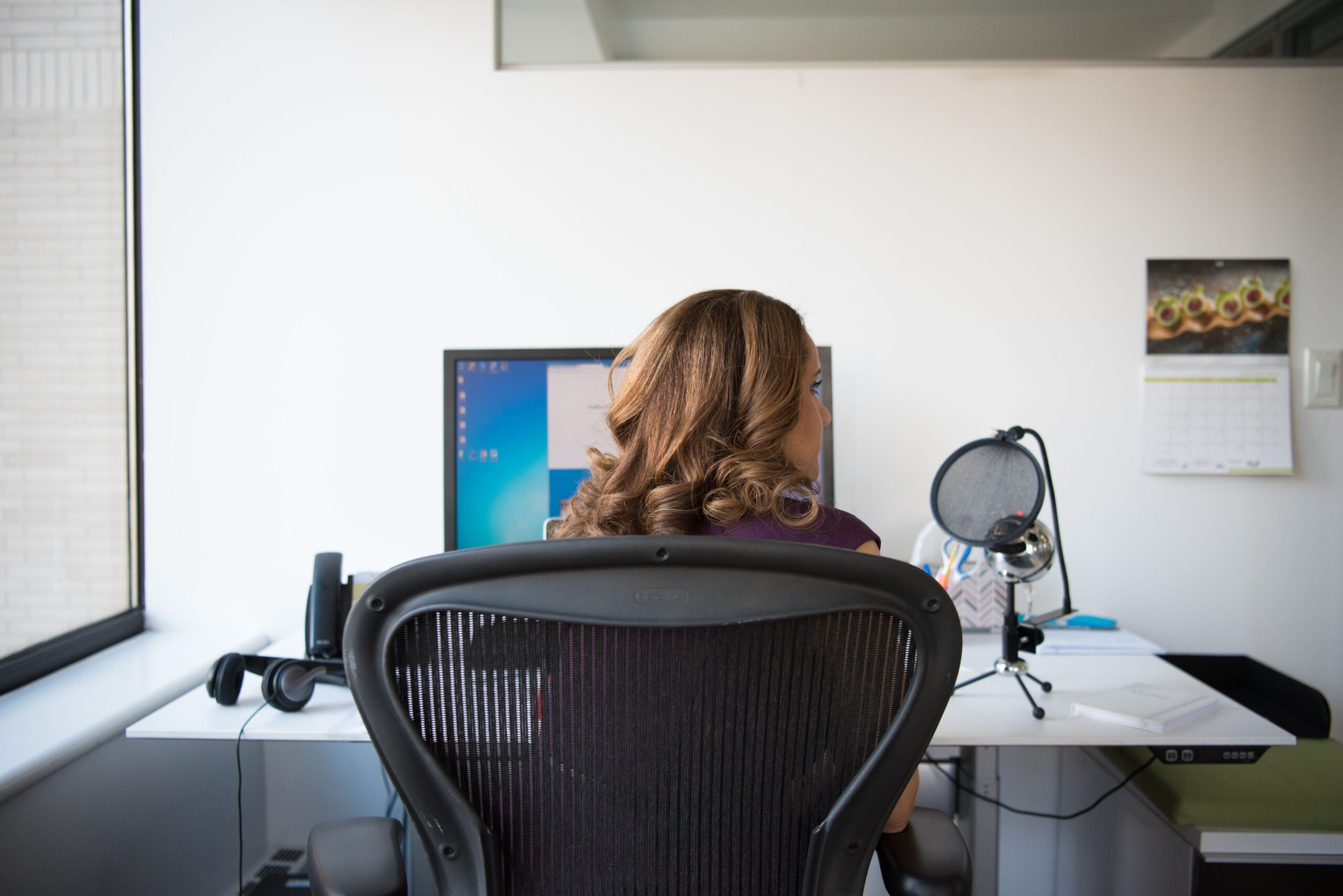 Woman sitting in an ergonomic mesh office chair at a home office desk with monitor and microphone